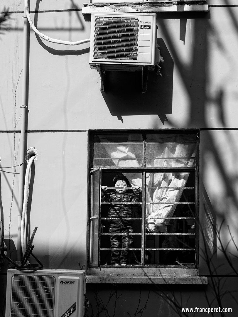 Kids behind the fence. I was attracted by 2 common point:<br />  1- the round face of the kid and the round shape of the air conditioner fan on top of him.<br /> 2 - His straight and symmetric posture compare to the straight lines (tube, pipe, fence, window, shadows) surrounding him.